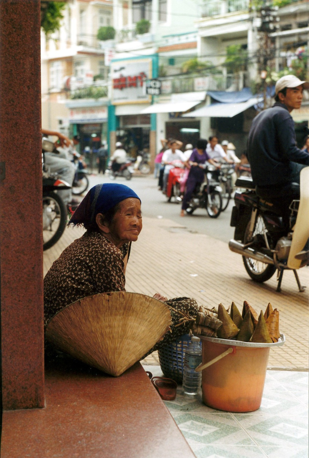 Cụ bà bán bánh trên phố đông người qua lại.
