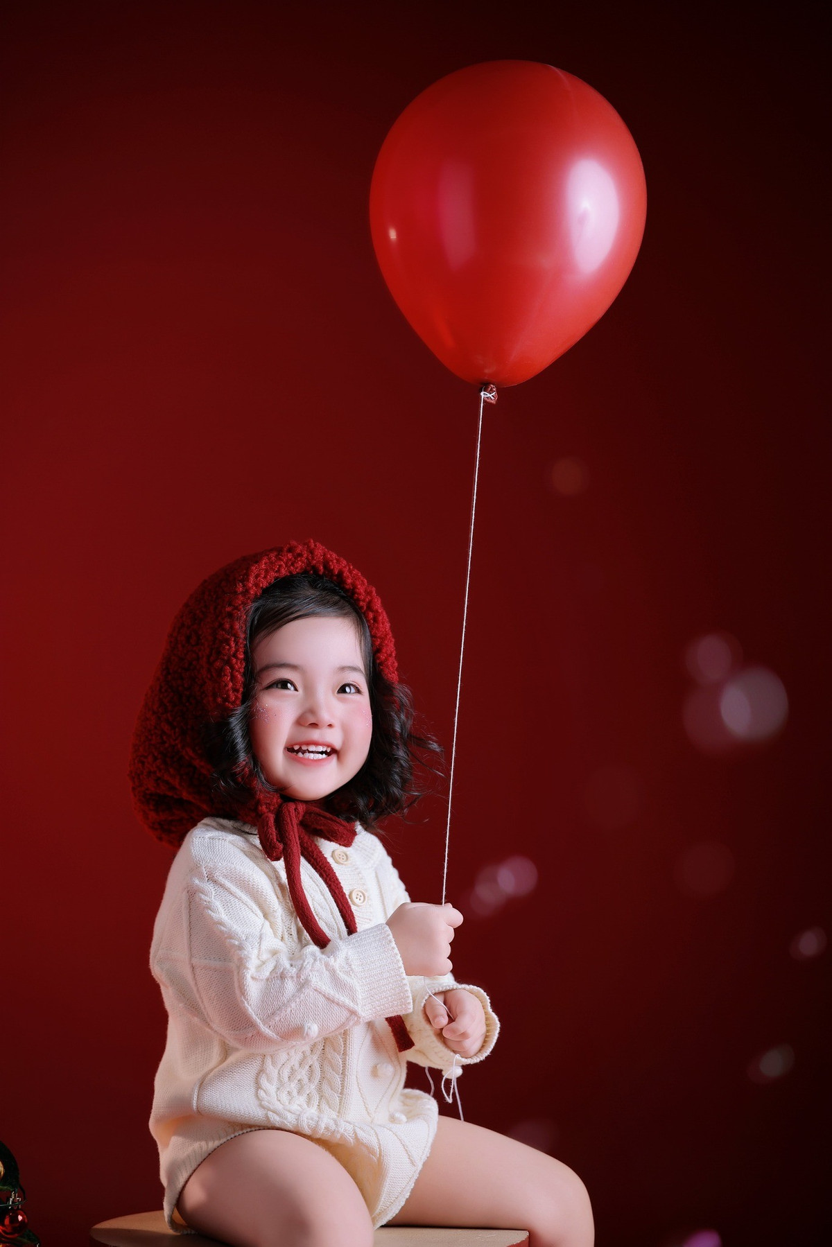 Cô bé diện áo len trắng dáng rộng, kết hợp cùng mũ len đỏ xinh xắn, tay cầm quả bóng bay đỏ nổi bật trên phông nền cùng tông màu.