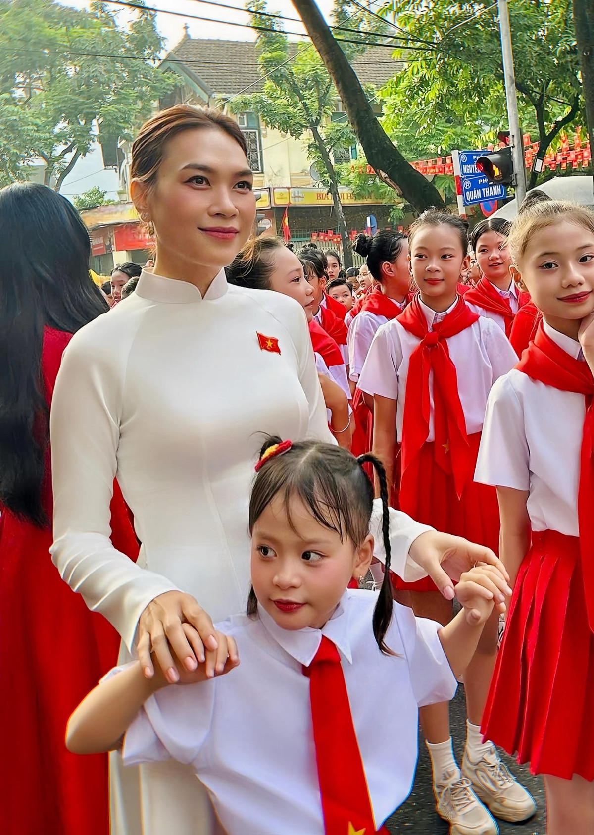 Không diện trang phục cầu kỳ, Mỹ Tâm chọn áo dài trắng tinh khôi, ôm dáng thanh thoát, điểm xuyết chiếc huy hiệu cờ đỏ sao vàng trang trọng. Tóc búi gọn sau gáy, gương mặt trang điểm nhẹ nhàng với đôi mắt sáng và bờ môi hồng tự nhiên, mang đến tổng thể vừa giản dị, vừa sang trọng.