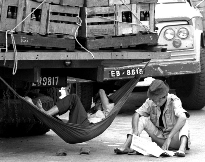 Một tài xế mắc võng ngủ trưa dưới gầm xe tải, cạnh đó là một người ngồi đọc báo, Sài Gòn năm 1970.