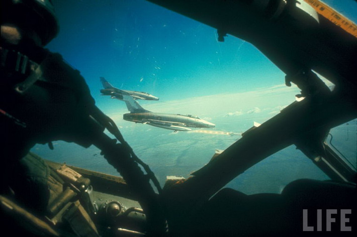 Bên ngoài cửa sổ buồng lái, một chiến đấu cơ F-100 của Mỹ đang phóng tên lửa Bullpup vào mục tiêu mặt đất ở miền Nam Việt Nam.