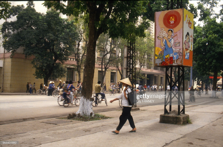 Trên vỉa hè bờ hồ Hoàn Kiếm, đối diện Bưu điện Hà Nội. Trên cột điện có tranh cổ động về Đại hội Đảng toàn quốc lần thứ VI, kỳ Đại hội đưa ra đường lối Đổi Mới cho đất nước.