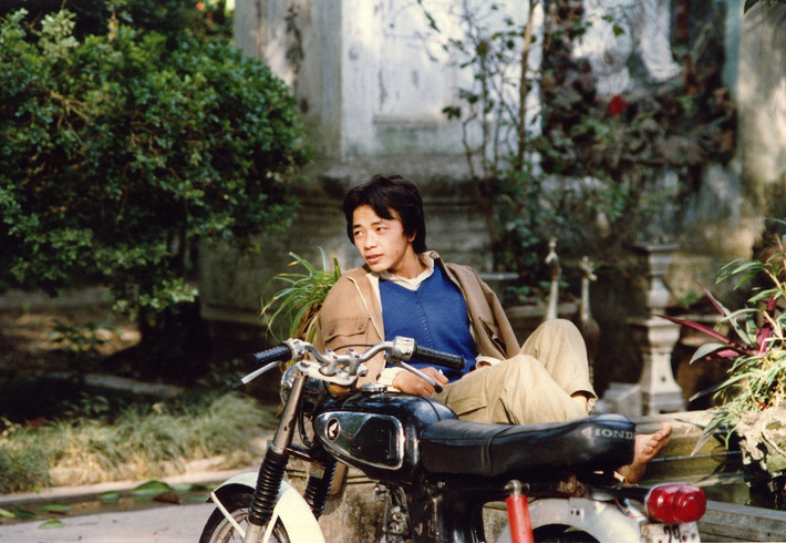 Người thanh niên nằm thảnh thơi trong khuôn viên một ngôi đền hoặc chùa, cạnh chiếc xe Honda 67 bóng loáng.