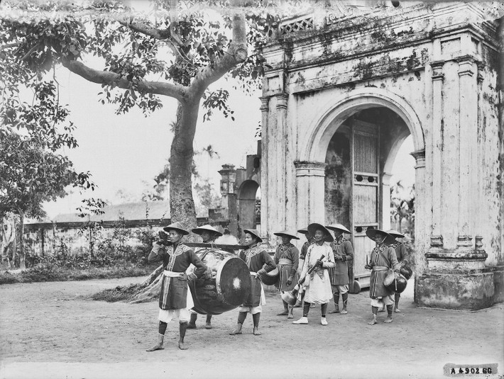 Người cầm loa và các nhạc công bên ngoài cổng dinh quan phủ ở làng Luật Trung, phủ Kiến Xương, nay thuộc hay xã Vũ Quý và Vũ Trung, huyện Kiến Xương.