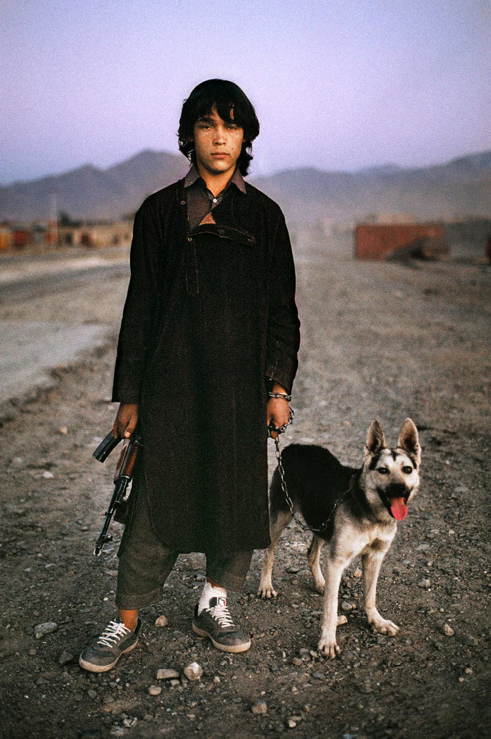 Cậu bé người Afghanistan dắt chó, tay cầm súng trường, đứng bên con đường dẫn đến Kandahar.