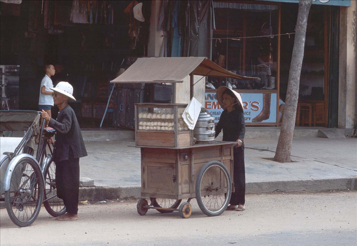 Cụ bà bán bánh bao dạo đẩy chiếc xe chở bánh của mình trên một con đường.