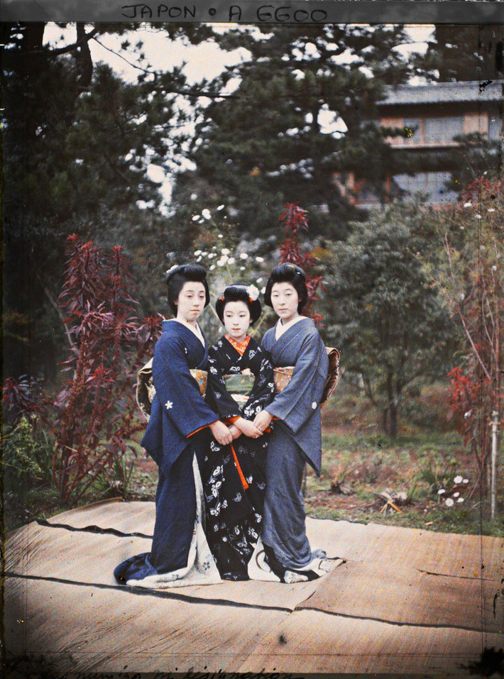 Hai geisha đứng bên một maiko (geisha tập sự) tại khu vườn của một okiya, nơi các geisa hành nghề, Kyoto, Nhật Bản năm 1912. Theo quy định của nghề geisha, các maiko mặc áo cổ đỏ, còn geisa mặc áo cổ trắng. Ảnh: Stéphane Passet/Collections.albert-kahn.hauts-de-seine.fr.