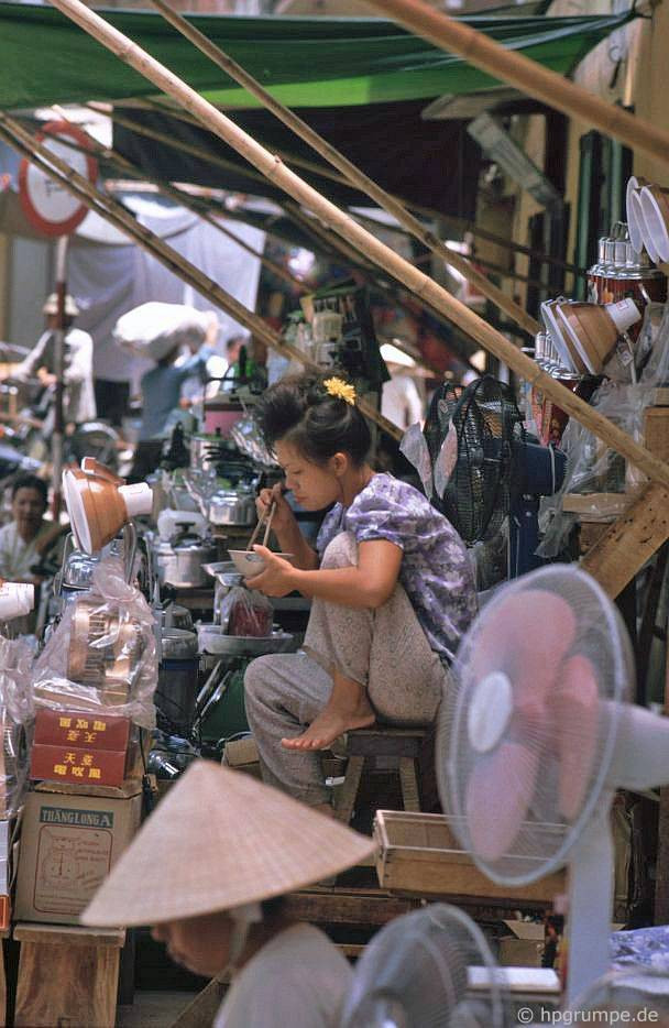 Người bánh hàng ngồi ăn trưa giữa đống hàng hóa gồm quạt điện, đèn bàn, nồi áp suất, phích nước...