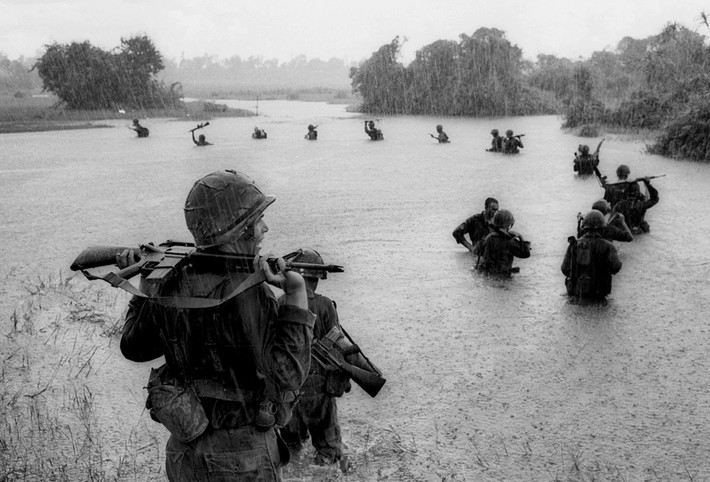 Lính dù của Tiểu Đoàn 2, Lữ đoàn dù 173 Mỹ giữ các khẩu súng tự động trên vai cho khỏi ướt khi vượt qua một con sông trong cơn mưa tại khu vực rừng Bến Cát, Sông Bé, vào ngày 25/9/1965. Ảnh: AP/Henri Huet.