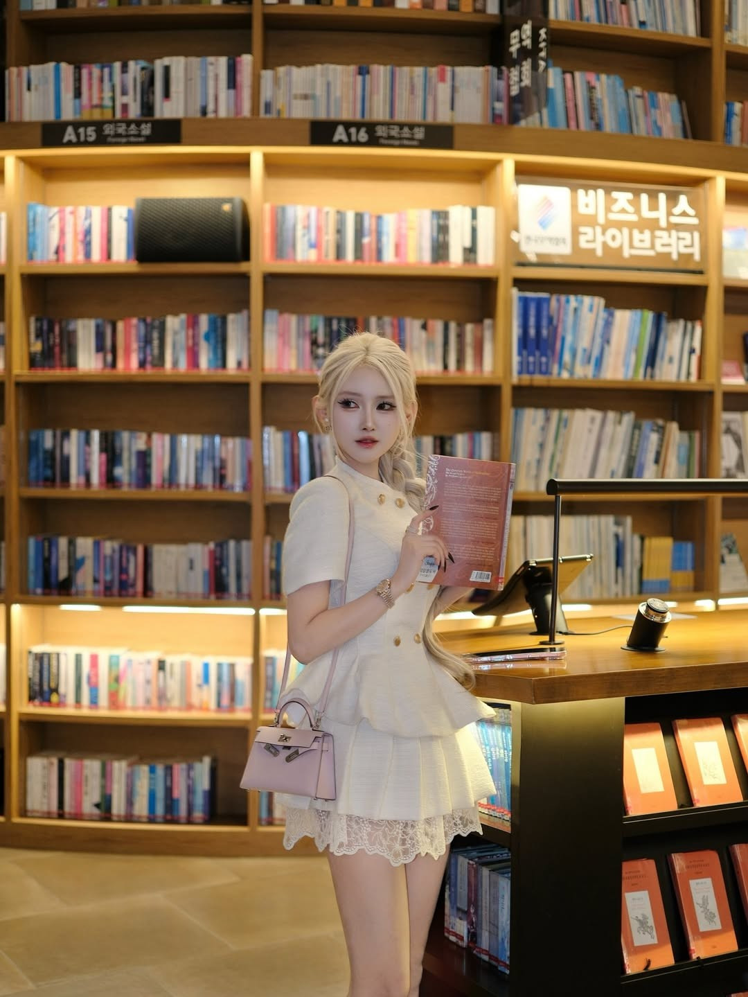 Mái tóc bạch kim được tết dài gọn gàng giúp tôn lên gương mặt nhỏ nhắn, làn da trắng mịn và đường nét thanh tú. Ánh mắt to tròn, sống mũi cao cùng đôi môi hồng chúm chím mang đến cảm giác mong manh, tựa một “búp bê sống” giữa những giá sách cao vút.