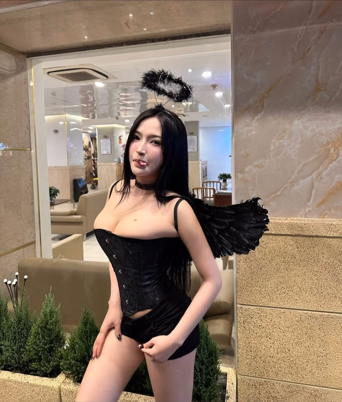 Chiếc corset ôm sát cơ thể tôn trọn đường cong hoàn hảo, kết hợp cùng đôi cánh đen và vòng halo lông vũ, khiến tổng thể vừa gợi cảm vừa đầy mê hoặc.