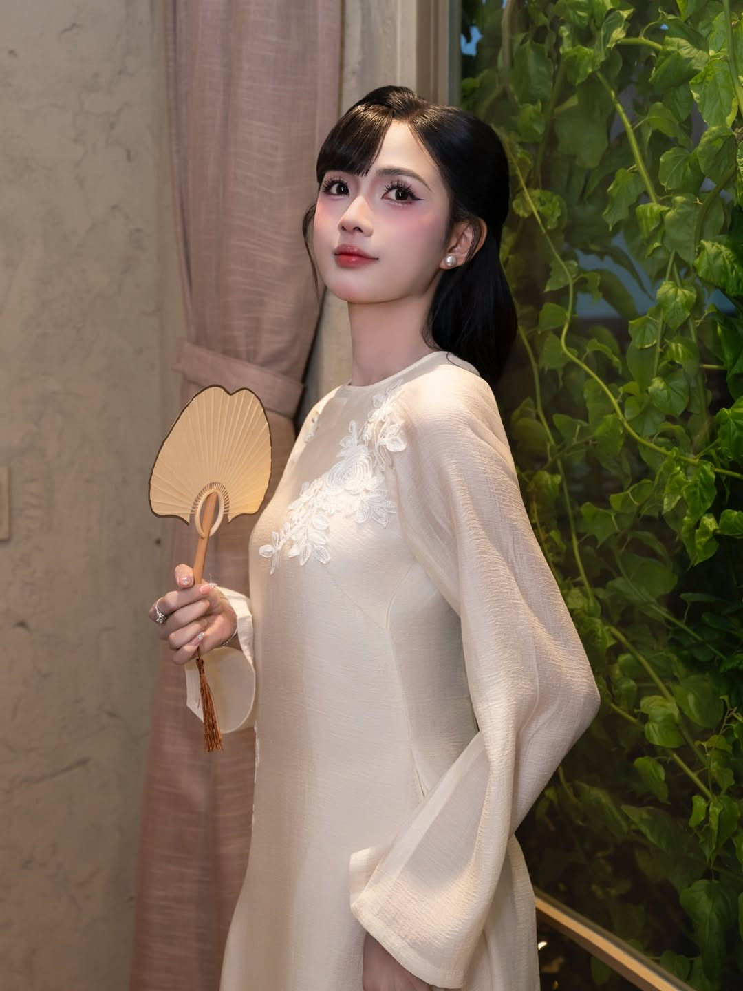 Mái tóc đen dài được buộc gọn phía sau, phần mái uốn cong ôm nhẹ gương mặt giúp tôn lên những đường nét thanh tú. Lối trang điểm trong trẻo, tập trung vào đôi mắt to tròn và làn da mịn màng càng làm nổi bật vẻ đẹp dịu dàng, nền nã của nữ tiếp viên hàng không. Phụ kiện đi kèm cũng được tiết chế tối đa: hoa tai ngọc trai nhỏ, chiếc quạt tay truyền thống và túi xách tông ấm tạo điểm nhấn vừa đủ mà không làm mất đi tinh thần của bộ áo dài.