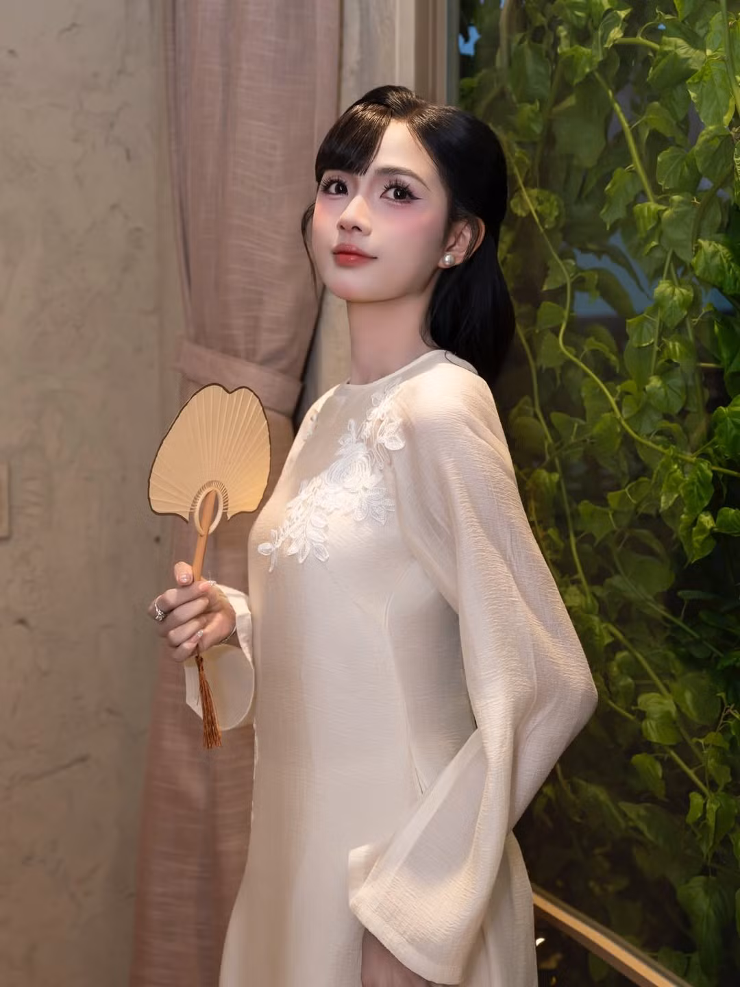 Mái tóc đen dài được buộc gọn phía sau, phần mái uốn cong ôm nhẹ gương mặt giúp tôn lên những đường nét thanh tú. Lối trang điểm trong trẻo, tập trung vào đôi mắt to tròn và làn da mịn màng càng làm nổi bật vẻ đẹp dịu dàng, nền nã của nữ tiếp viên hàng không. Phụ kiện đi kèm cũng được tiết chế tối đa: hoa tai ngọc trai nhỏ, chiếc quạt tay truyền thống và túi xách tông ấm tạo điểm nhấn vừa đủ mà không làm mất đi tinh thần của bộ áo dài.