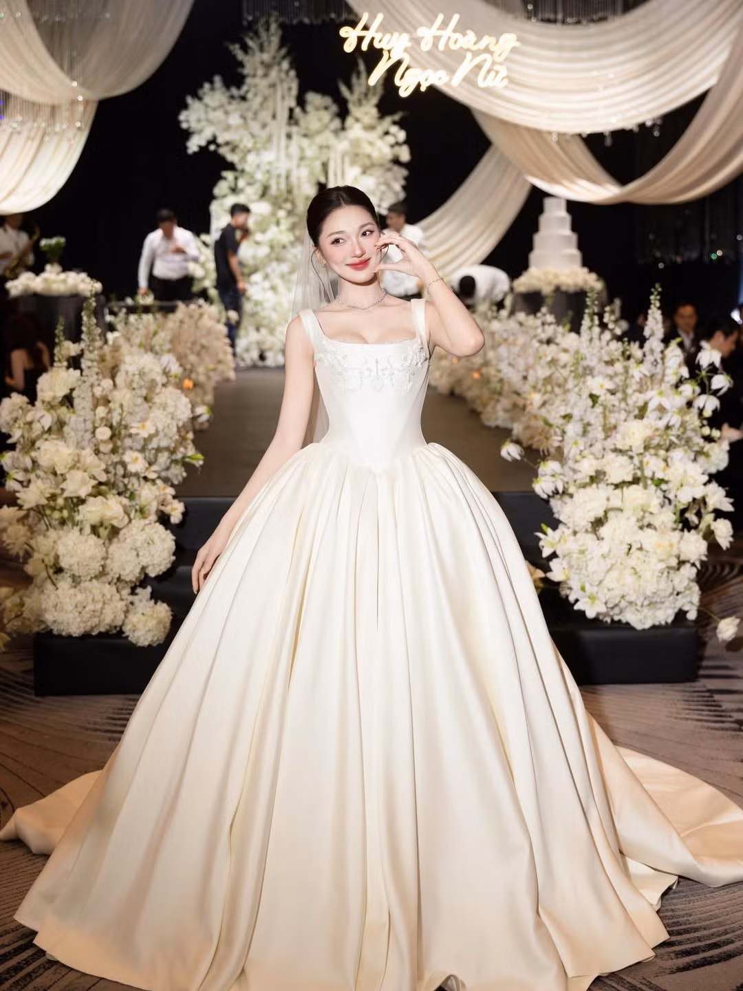 Xuất hiện trong không gian lễ cưới được trang hoàng lộng lẫy với hoa trắng và ánh đèn ấm áp, Nguyễn Ngọc Nữ khoác lên mình váy cưới phom dáng công chúa với phần thân trên ôm sát, tôn vòng eo thon gọn, kết hợp chân váy xòe bồng bề thế. Chất liệu lụa – satin cao cấp cùng những chi tiết thêu đính tinh xảo giúp tổng thể bộ váy vừa thanh lịch vừa mang hơi thở cổ tích.
