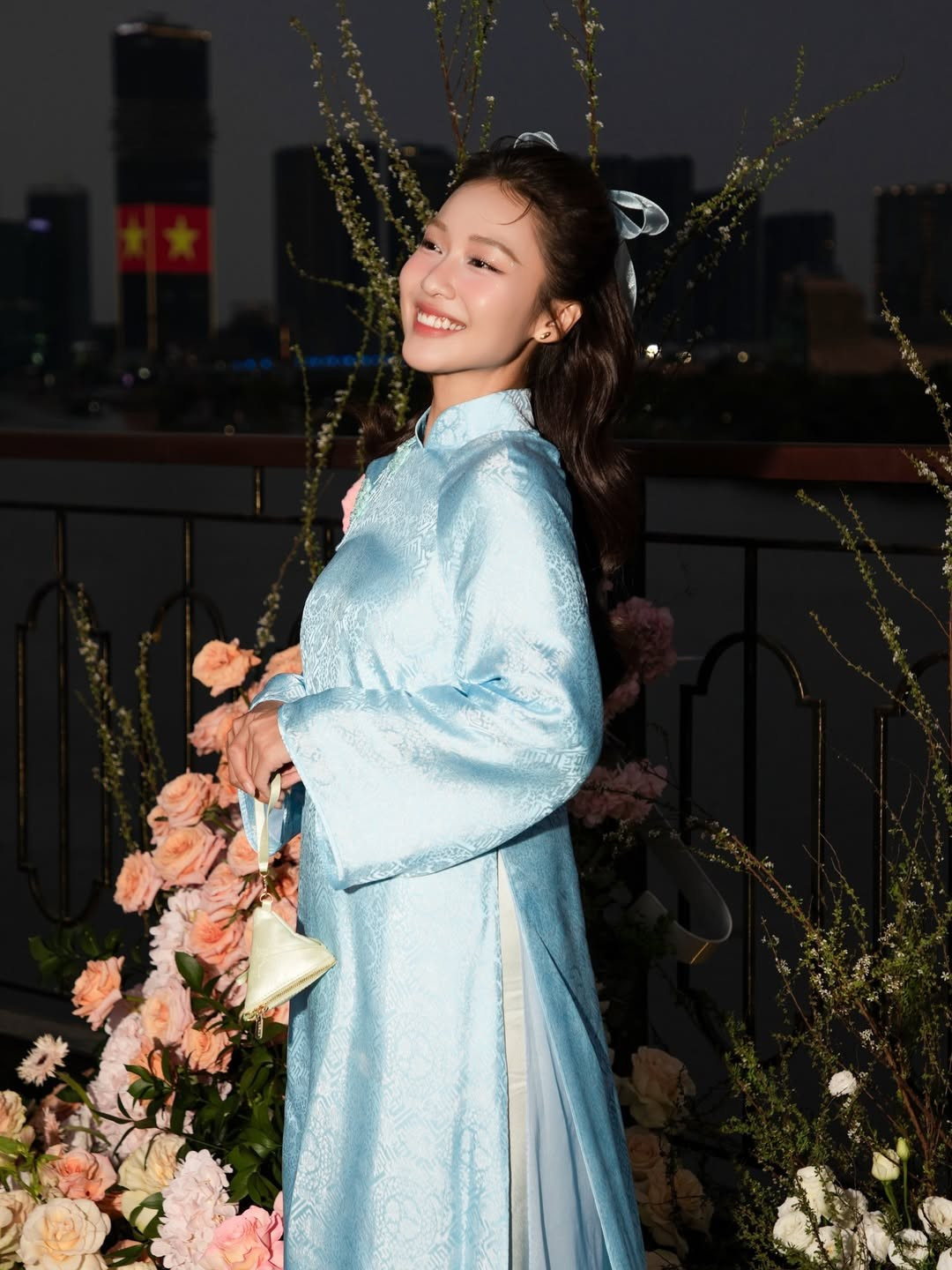 Không chỉ ghi dấu ấn trong không gian trong nhà, Khả Ngân còn thực hiện nhiều khung hình ngoài trời khi hoàng hôn buông xuống. Giữa phông nền thành phố lên đèn và những cụm hoa trang trí rực rỡ, tà áo dài xanh càng trở nên nổi bật. Sự đối lập giữa vẻ đẹp truyền thống của trang phục và nhịp sống đô thị hiện đại tạo nên tổng thể hình ảnh đầy tính nghệ thuật.