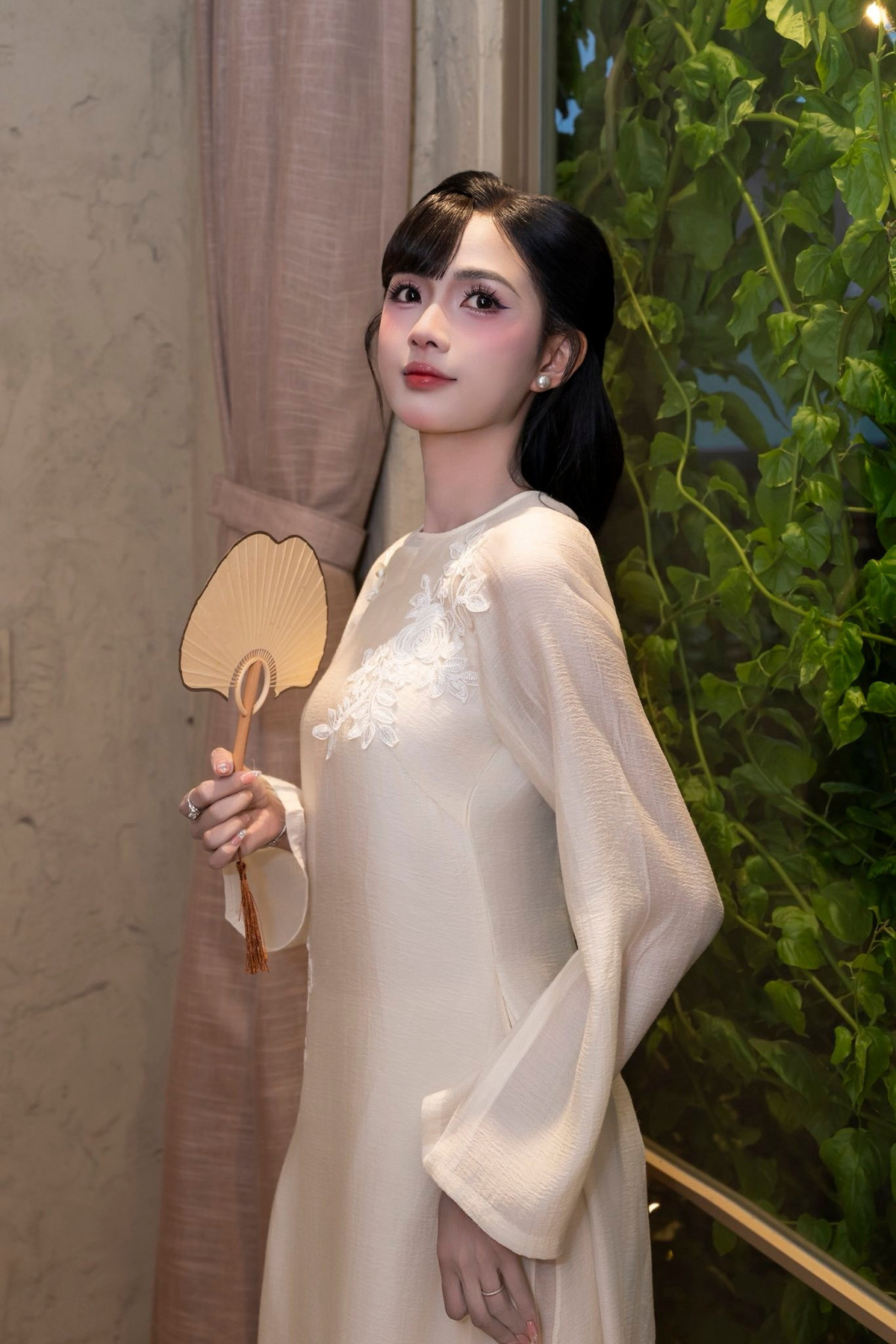 Điểm nhấn đặc biệt trong tạo hình lần này chính là mái tóc được búi gọn sau gáy, điểm xuyết bông hoa trắng tinh khôi. Kiểu tóc này giúp tôn lên gương mặt thanh tú cùng đường nét mềm mại của cô. Lối trang điểm nhẹ nhàng với tông hồng đất, đôi môi đỏ vừa phải và làn da mịn màng giúp tổng thể thêm phần hài hòa, đúng tinh thần “đẹp mà không cần phô trương”.