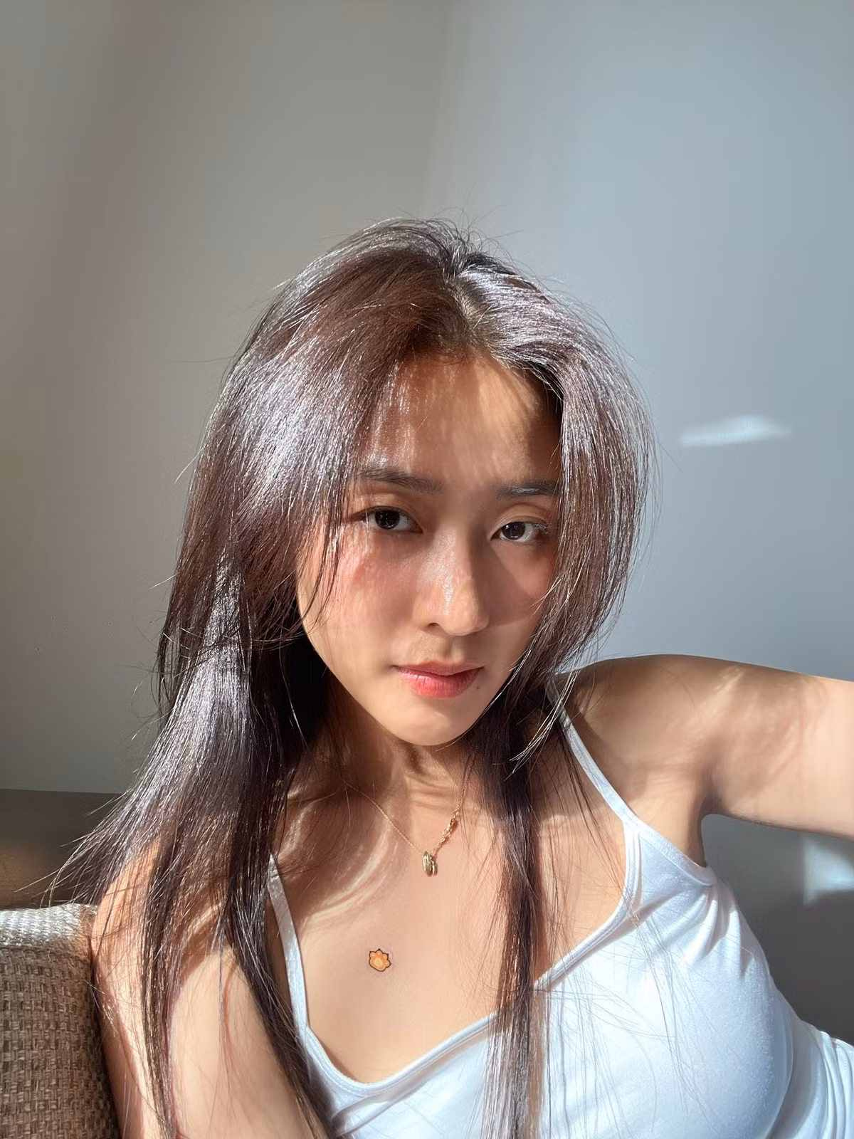 Ánh nắng chiếu xuyên qua cửa sổ tạo nên những mảng sáng – tối mềm mại trên gương mặt, tôn lên làn da căng bóng, đôi mắt sâu và sống mũi thanh thoát. Mái tóc suôn dài thả tự nhiên càng làm tăng nét nữ tính và trẻ trung.