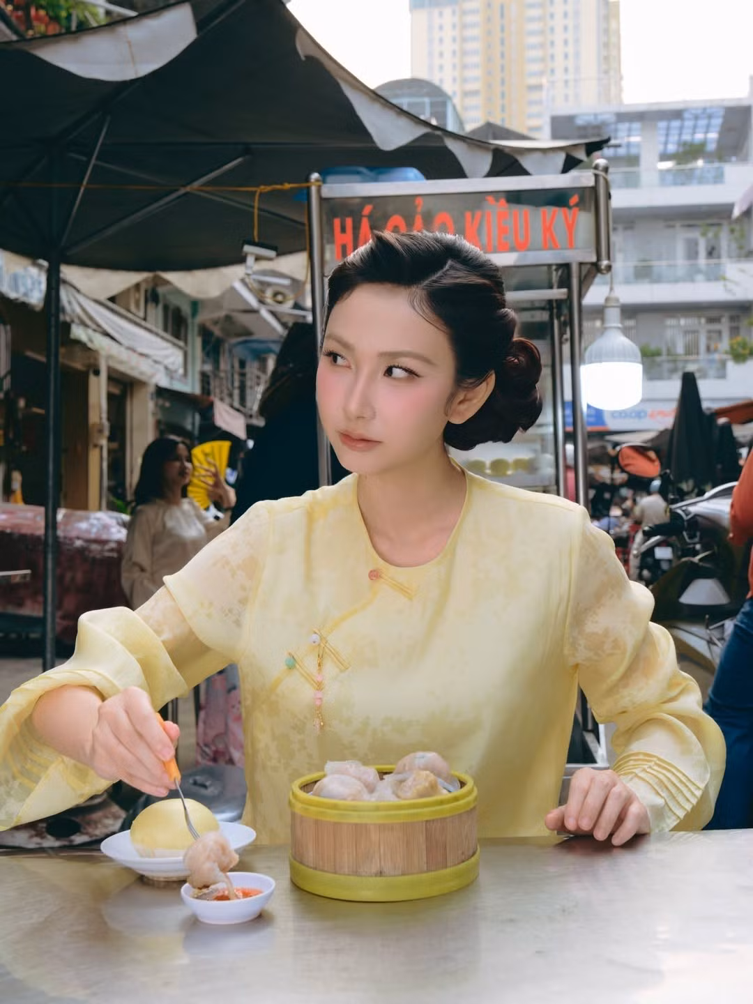 Ngồi trước xửng há cảo nóng hổi, Thanh Mèo thoải mái thưởng thức món ăn dân dã, tạo nên hình ảnh đối lập thú vị giữa tà áo dài duyên dáng và không khí đời thường, tấp nập phía sau.