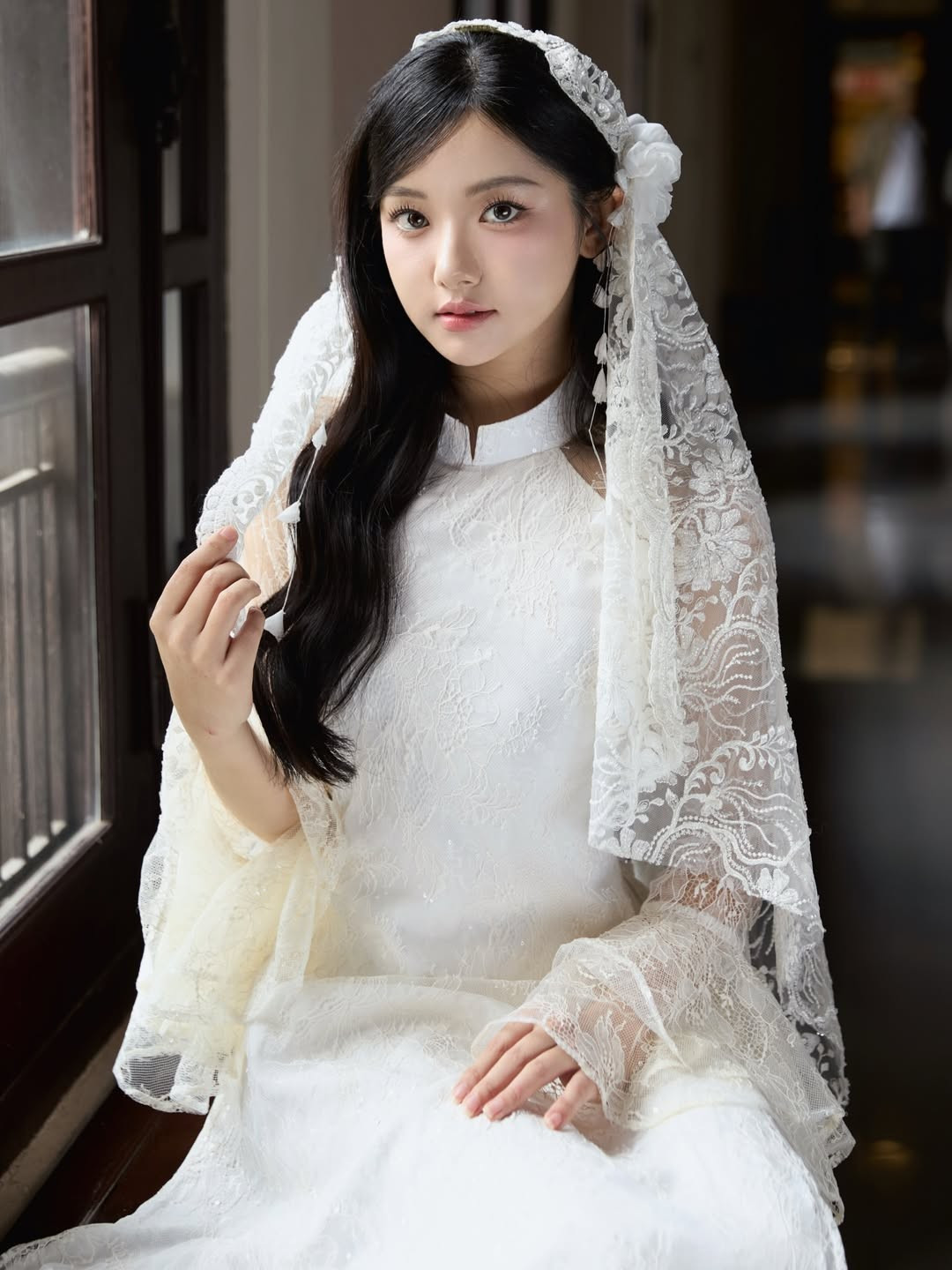 Chiếc áo dài trắng được thiết kế từ chất liệu ren mỏng, tạo hiệu ứng xuyên sáng nhẹ nhàng nhưng vẫn giữ được nét kín đáo cần có. Họa tiết ren hoa chạy dọc thân áo và phần tay áo dài giúp tổng thể thêm phần mềm mại, bay bổng. Phom dáng ôm vừa vặn tôn lên vóc dáng thanh mảnh, trong khi phần cổ cao giữ trọn tinh thần truyền thống của trang phục.