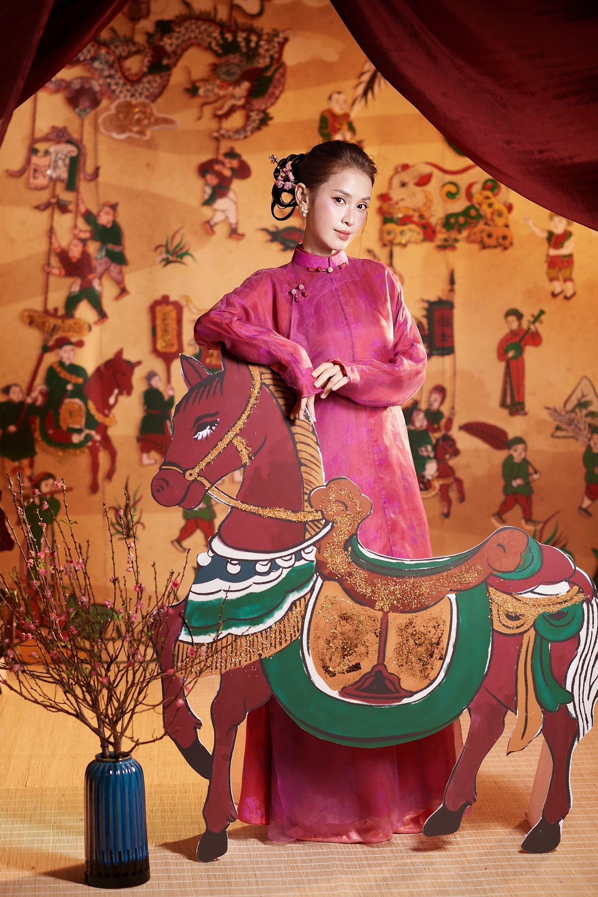Không gian chụp ảnh cũng góp phần làm nổi bật thần thái của Lê Tú Vi. Phông nền lấy cảm hứng từ tranh dân gian với hình ảnh rồng, ngựa, nhân vật truyền thống cùng sắc vàng ấm áp tạo nên bầu không khí đậm chất lễ hội. Những cành hoa đào được bố trí khéo léo vừa tạo chiều sâu cho khung hình, vừa tôn lên vẻ mềm mại của tà áo dài.