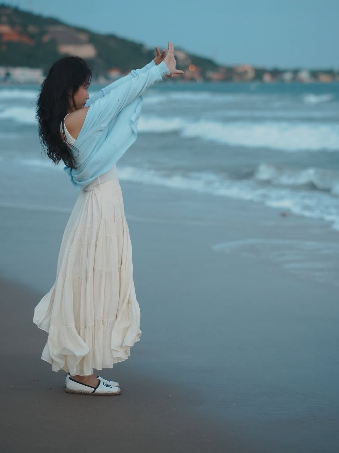 Trong khung cảnh biển chiều lộng gió, Khả Ngân lựa chọn trang phục đơn giản với váy trắng dáng dài kết hợp cardigan xanh pastel. Cách phối đồ tối giản nhưng tinh tế giúp cô tôn lên sắc vóc thon gọn, vóc dáng cân đối cùng thần thái dịu dàng. Mái tóc dài buông tự nhiên theo gió, từng chuyển động nhẹ cũng đủ tạo nên những khoảnh khắc đầy chất thơ, khiến tổng thể bức ảnh mang màu sắc điện ảnh, trầm lắng đúng chất cuối năm.