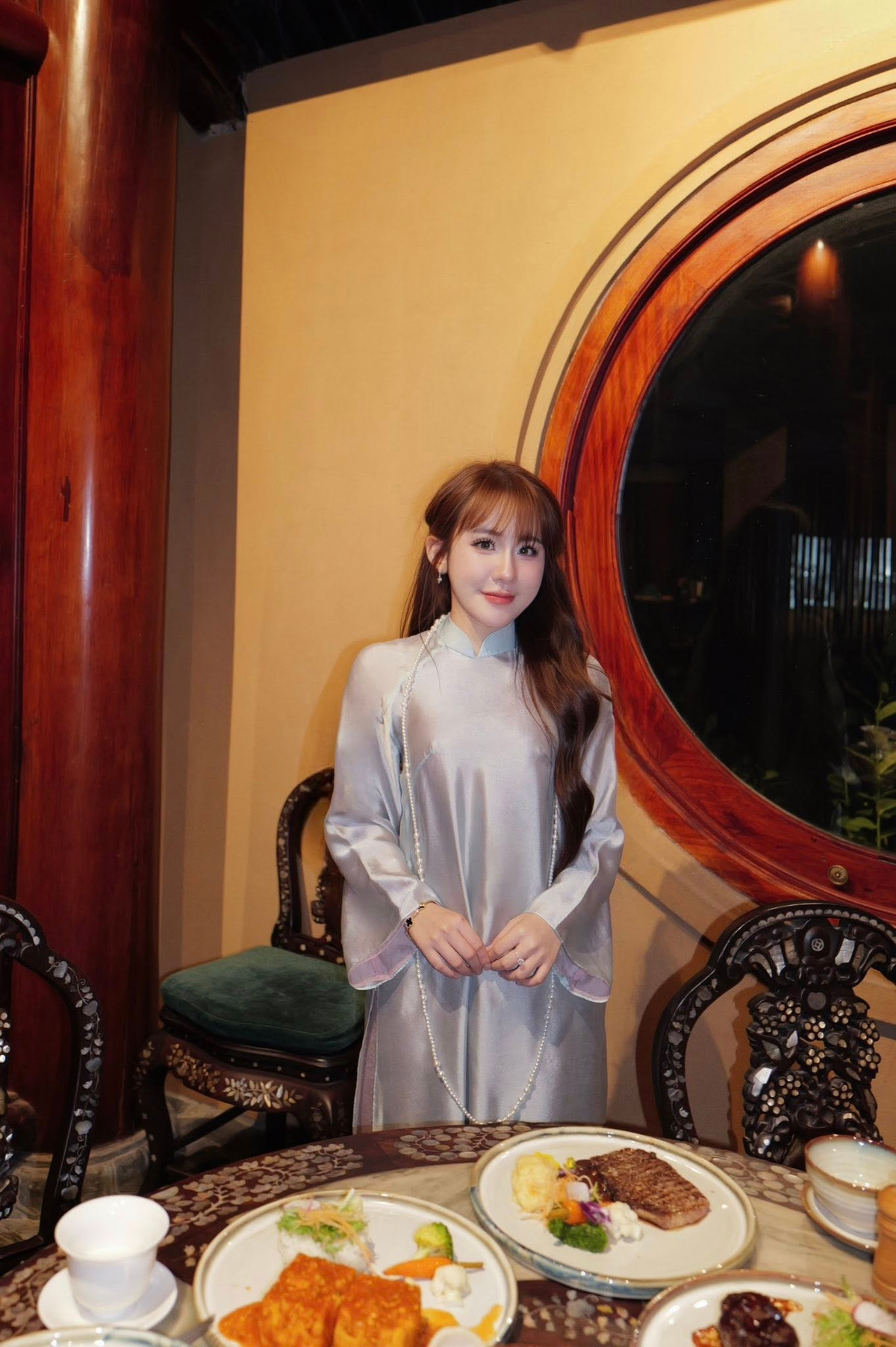 Bối cảnh chụp mang hơi hướng cổ điển với không gian gỗ chạm khắc tinh xảo, bàn trà và các món ăn được bày biện trang nhã. Sự kết hợp giữa trang phục áo dài và không gian truyền thống tạo nên tổng thể hài hòa, giúp bộ ảnh vừa sang trọng vừa gần gũi.