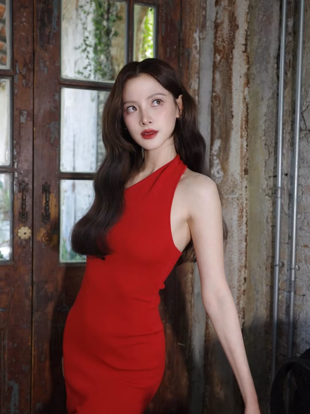 Làn da trắng mịn càng được tôn lên dưới sắc đỏ nổi bật của chiếc váy ôm sát, thiết kế lệch vai giúp khoe trọn bờ vai thon và phần cổ dài quyến rũ. Mái tóc dài uốn sóng nhẹ nhàng buông xuống làm tăng thêm vẻ mềm mại, sang trọng, tạo cảm giác vừa kiêu sa vừa thời thượng.