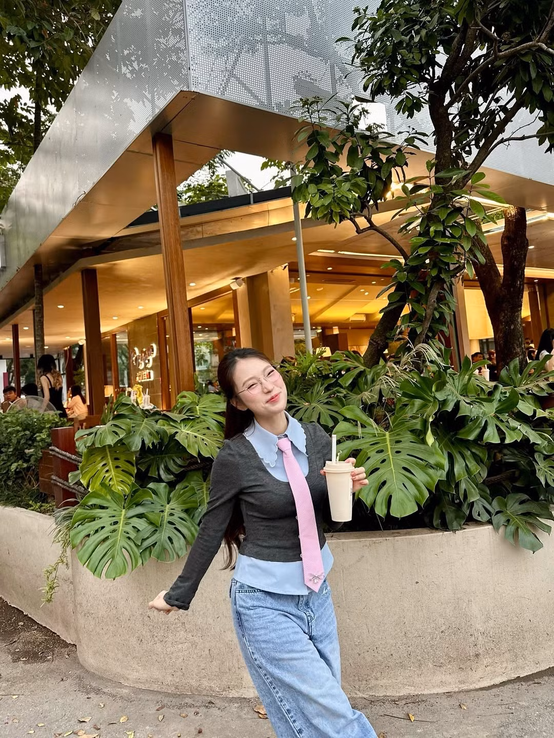 Trong loạt ảnh, Khánh Vy diện outfit đơn giản nhưng cực kỳ dễ thương với áo sơ mi xanh nhạt phối viền cổ bầu cách điệu. Cà vạt hồng pastel tạo điểm nhấn trẻ trung, tinh nghịch. Bên cạnh đó, quần jeans ống rộng mang hơi hướng retro, vừa thoải mái vừa hợp bối cảnh café ngoài trời.