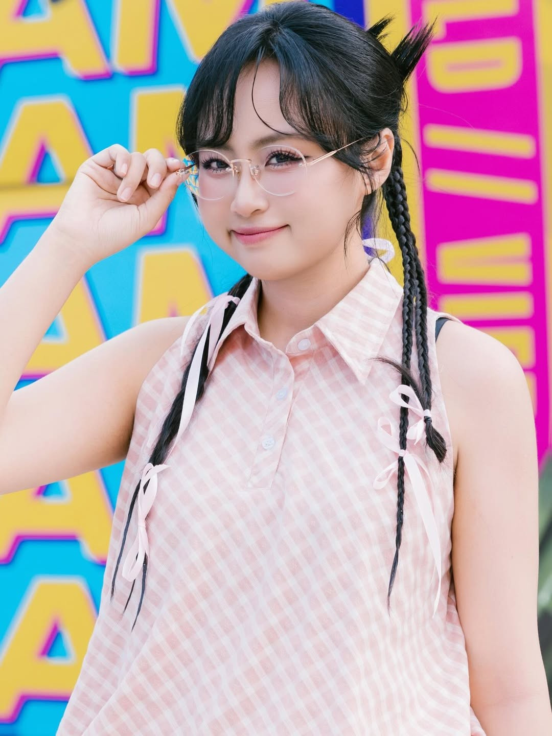 Kiểu tóc thắt bím hai bên giúp Minh Nghi thêm phần đáng yêu, tạo cảm giác như một cô nàng teen đầy năng lượng. Đôi kính trong suốt càng làm nổi bật vẻ ngây thơ, tươi sáng.