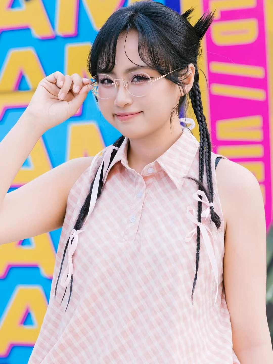 Kiểu tóc thắt bím hai bên giúp Minh Nghi thêm phần đáng yêu, tạo cảm giác như một cô nàng teen đầy năng lượng. Đôi kính trong suốt càng làm nổi bật vẻ ngây thơ, tươi sáng.