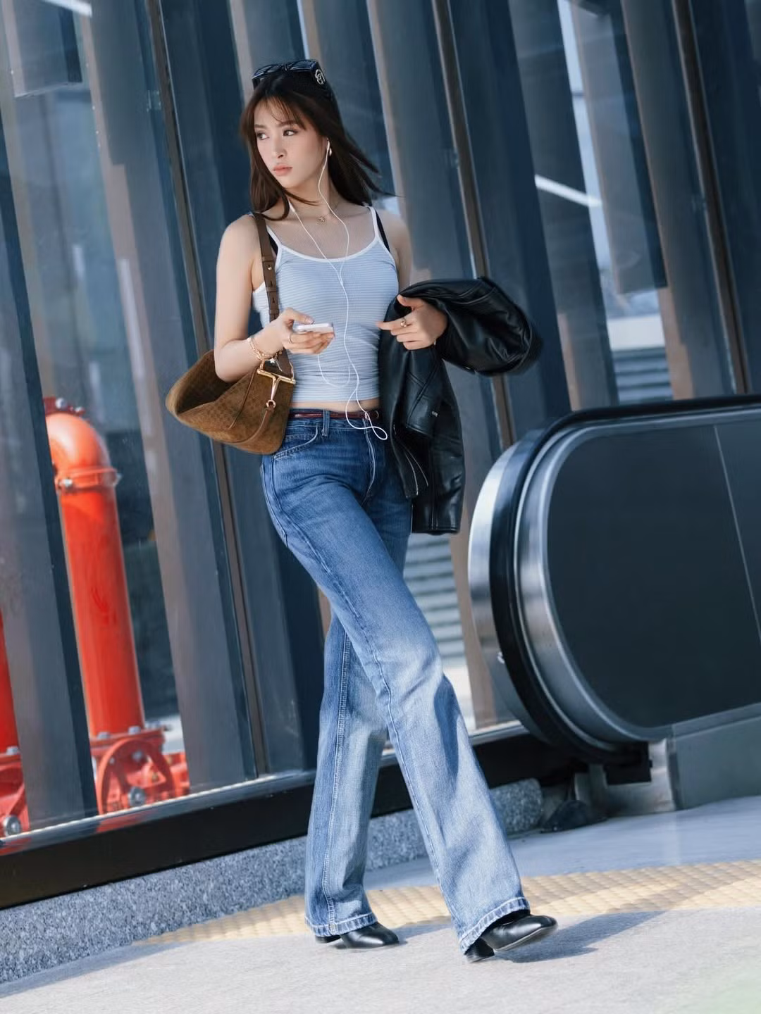 Trong loạt ảnh, Trần Tiểu Vy xuất hiện với set đồ street style năng động gồm áo hai dây dáng ôm tôn vòng eo thon gọn, quần jeans ống suông trẻ trung, phối cùng túi xách và áo khoác da khoác tay. Tai nghe trắng, kính mát cài đầu và mái tóc dài bay nhẹ trong gió tạo nên tổng thể vừa hiện đại, vừa phóng khoáng. Phong cách đời thường giản dị nhưng lại giúp nàng hậu khoe trọn vóc dáng cân đối cùng thần thái tự tin khi sải bước giữa không gian đô thị.