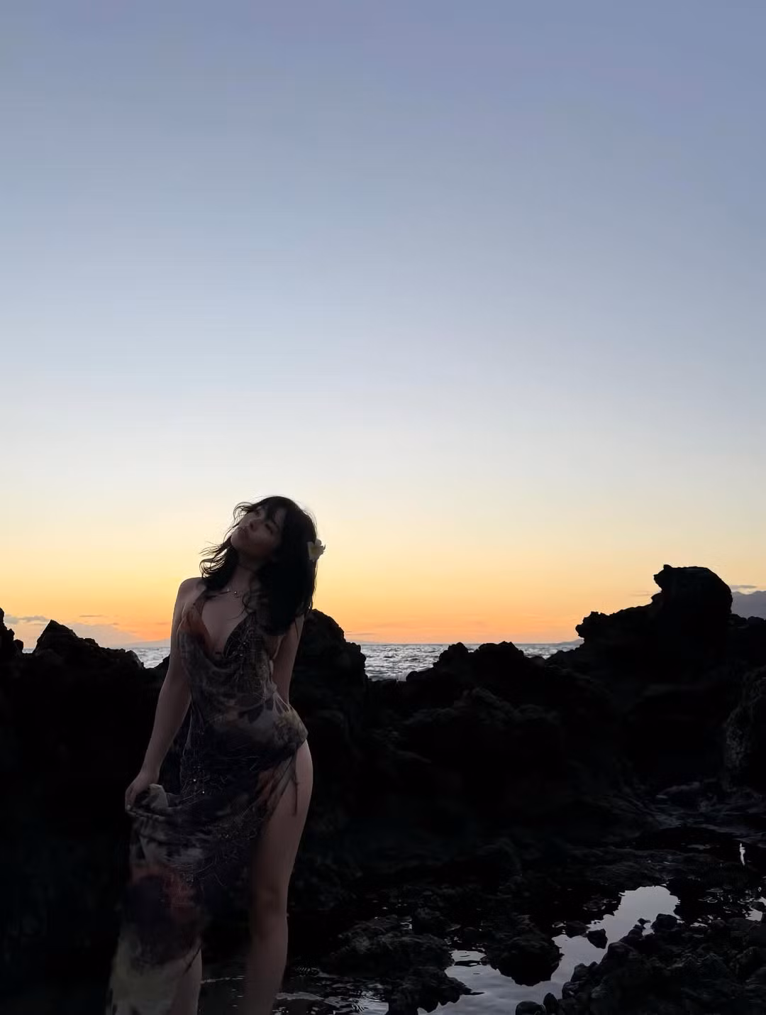 Việc sử dụng ánh sáng dạng tự nhiên từ mặt trời lặn đã tạo nên hiệu ứng viền sáng hoàn hảo, làm nổi bật mái tóc đen dài và đóa hoa trắng cài trên tóc cô, mang lại vẻ đẹp vừa hoang dại, vừa lãng mạn.