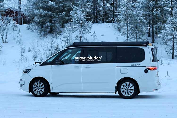 Trong ảnh, chiếc xe Volkswagen Multivan T7 2027 nguyên mẫu được ngụy trang với các hình dán Ocean trên phần thân sau, đang được thử nghiệm trong điều kiện thời tiết lạnh giá ở Thụy Điển với đèn pha hoàn toàn mới, lớn hơn một chút so với đèn pha của mẫu California tiền nhiệm.