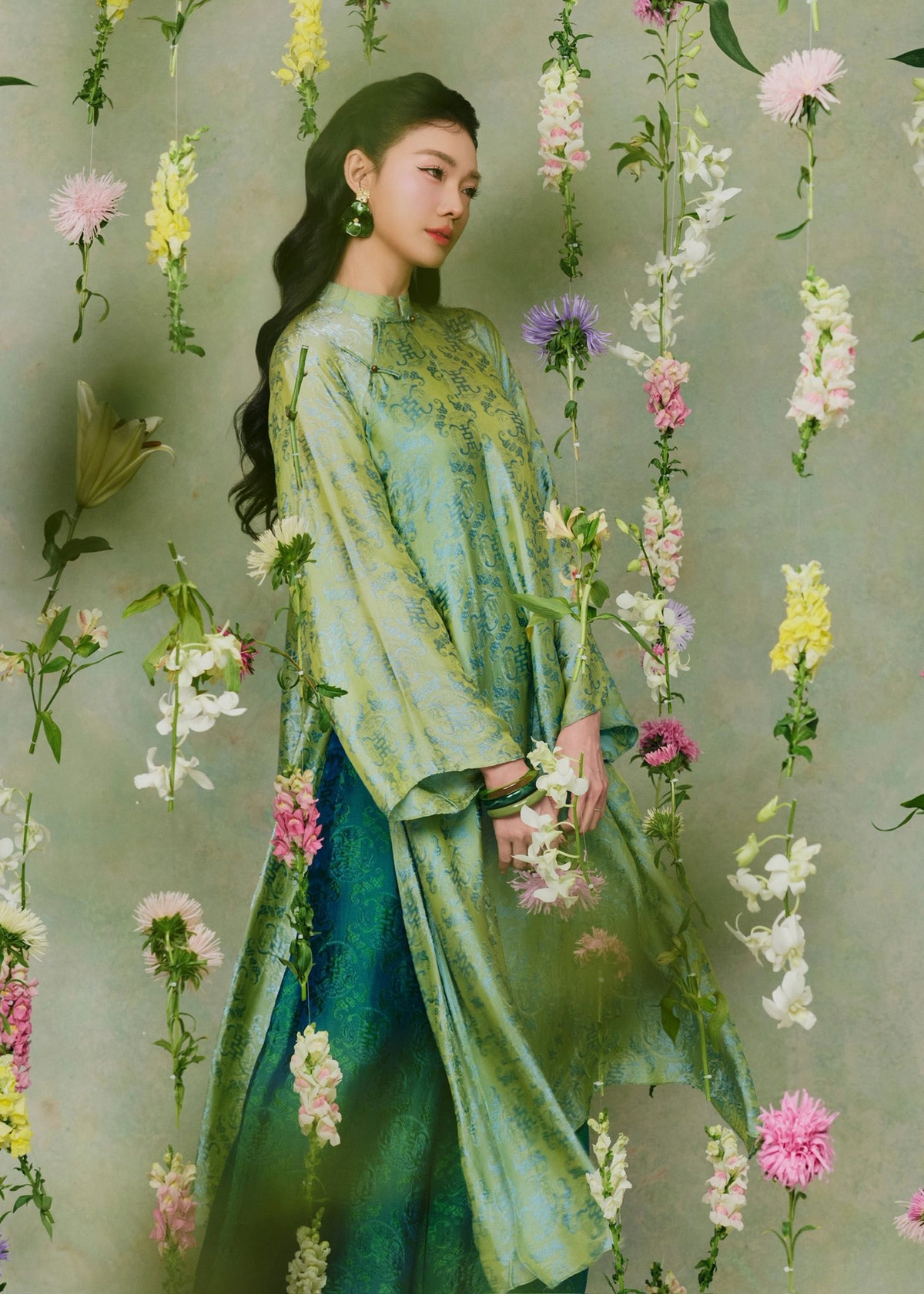 Thiết kế sử dụng chất liệu gấm cao cấp với họa tiết cổ điển chìm, tạo hiệu ứng thị giác sang trọng mà vẫn giữ được nét mềm mại đặc trưng của tà áo dài Việt. Gam màu xanh ngọc pha ánh vàng nhẹ được xem là điểm nhấn đắt giá, vừa tôn làn da sáng vừa gợi cảm giác thanh khiết, trang nhã.