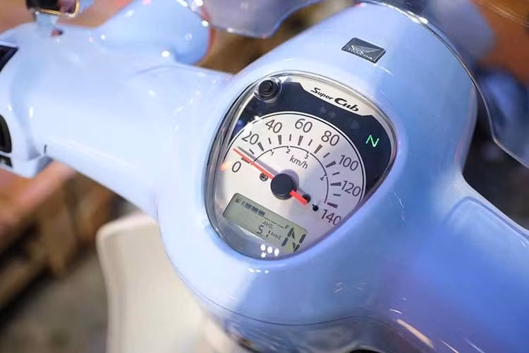 Honda Super Cub Fujisan 110 2026 vẫn sử dụng chìa khóa dạng cơ cổ điển. Đồng hồ tốc độ analog kèm màn hình LCD cỡ nhỏ, các nút bấm đơn giản không thay đổi. Bộ tem phối ba màu xanh dương nhạt, đậm và đỏ trên dàn áo phía trước.