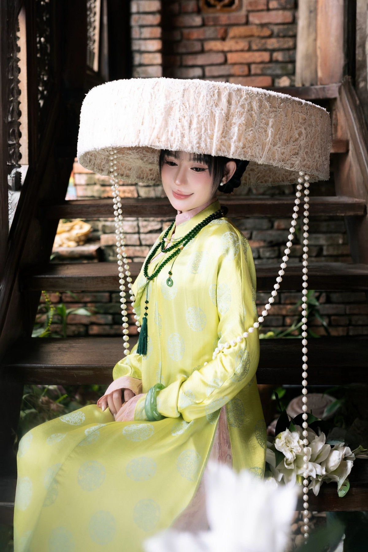 Phom dáng áo dài suông, tay dài, cổ cao vừa vặn giúp tôn lên vóc dáng thanh mảnh, đồng thời giữ được vẻ kín đáo, trang nhã đúng tinh thần trang phục truyền thống. Điểm nhấn đặc biệt là chiếc mấn đội đầu cỡ lớn, kết hợp chuỗi ngọc trai buông nhẹ hai bên, tạo nên tổng thể mang đậm phong vị xưa.