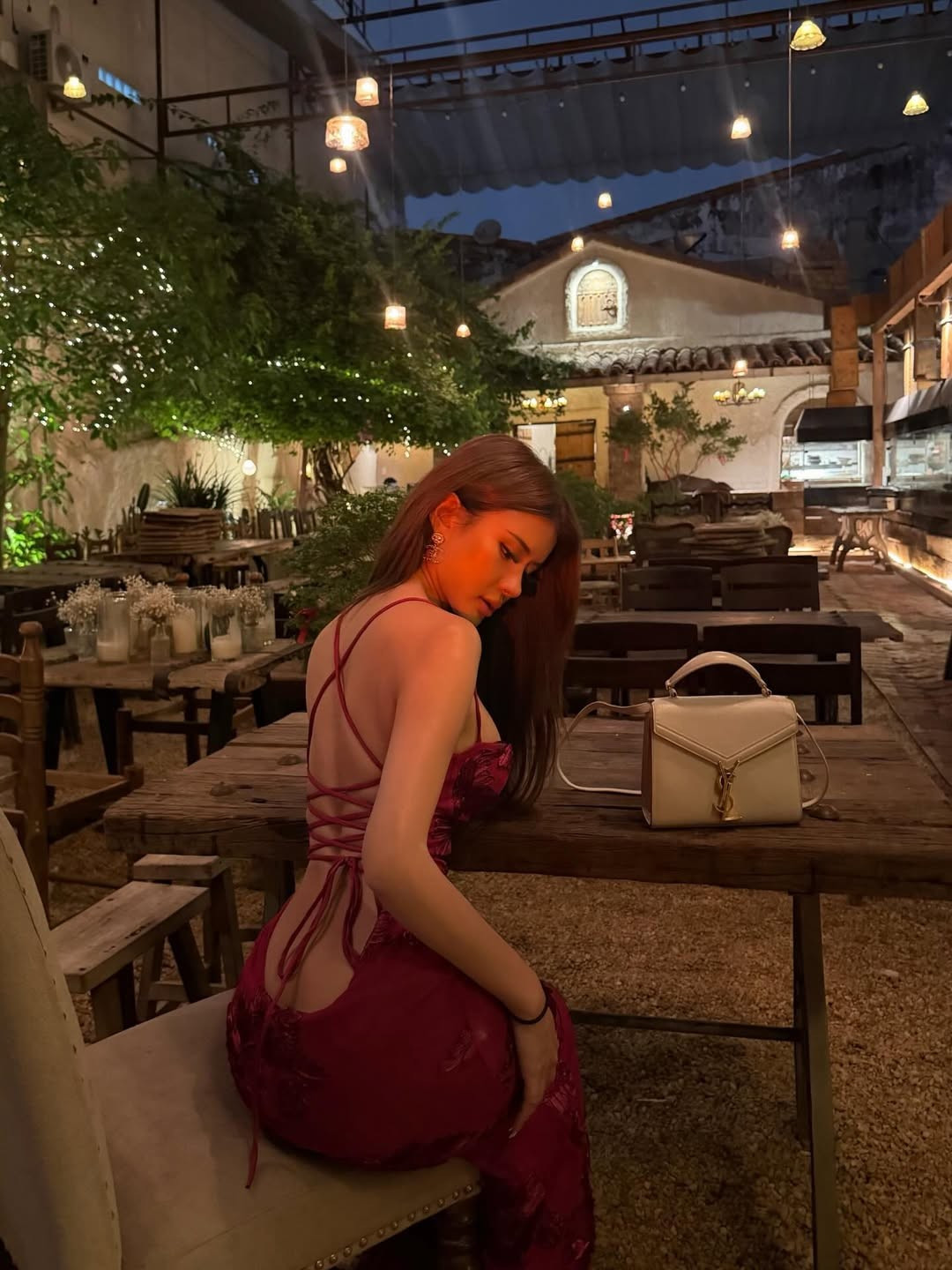 Bối cảnh chụp hình được lựa chọn kỹ lưỡng với không gian mang hơi hướng cổ điển, bàn ghế gỗ và ánh đèn vàng tạo cảm giác ấm áp. Sự kết hợp giữa sắc đỏ nổi bật của trang phục và tông màu trầm của background mang lại hiệu ứng thị giác mạnh mẽ, khiến nhân vật chính càng thêm nổi bật. Chiếc túi xách gam màu trung tính đi kèm cũng giúp tổng thể trở nên hài hòa, sang trọng.