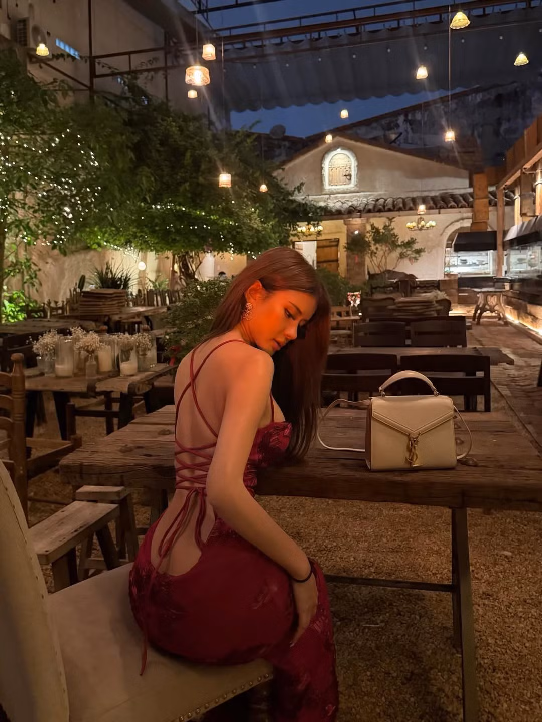 Bối cảnh chụp hình được lựa chọn kỹ lưỡng với không gian mang hơi hướng cổ điển, bàn ghế gỗ và ánh đèn vàng tạo cảm giác ấm áp. Sự kết hợp giữa sắc đỏ nổi bật của trang phục và tông màu trầm của background mang lại hiệu ứng thị giác mạnh mẽ, khiến nhân vật chính càng thêm nổi bật. Chiếc túi xách gam màu trung tính đi kèm cũng giúp tổng thể trở nên hài hòa, sang trọng.