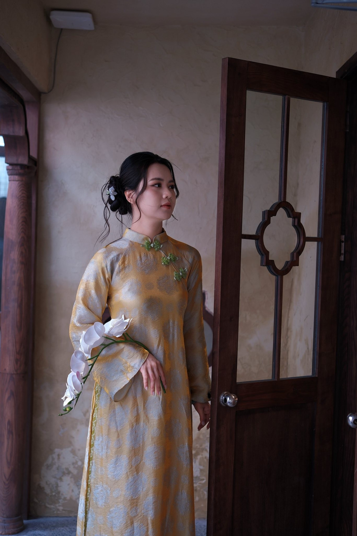 Không gian chụp mang phong cách truyền thống với ánh đèn vàng ấm, nội thất gỗ và bàn trà đậm chất Á Đông. Tổng thể khung hình gợi cảm giác hoài niệm, trang nhã, làm nổi bật hình ảnh người phụ nữ Việt Nam dịu dàng trong tà áo dài.