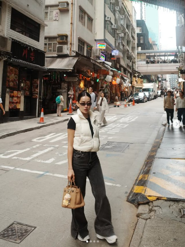 Xuất hiện trên đường phố Hong Kong, Milan Phạm lựa chọn phong cách casual-chic với áo thun tối màu kết hợp áo gile phao trắng, quần jeans ống rộng và sneakers năng động.