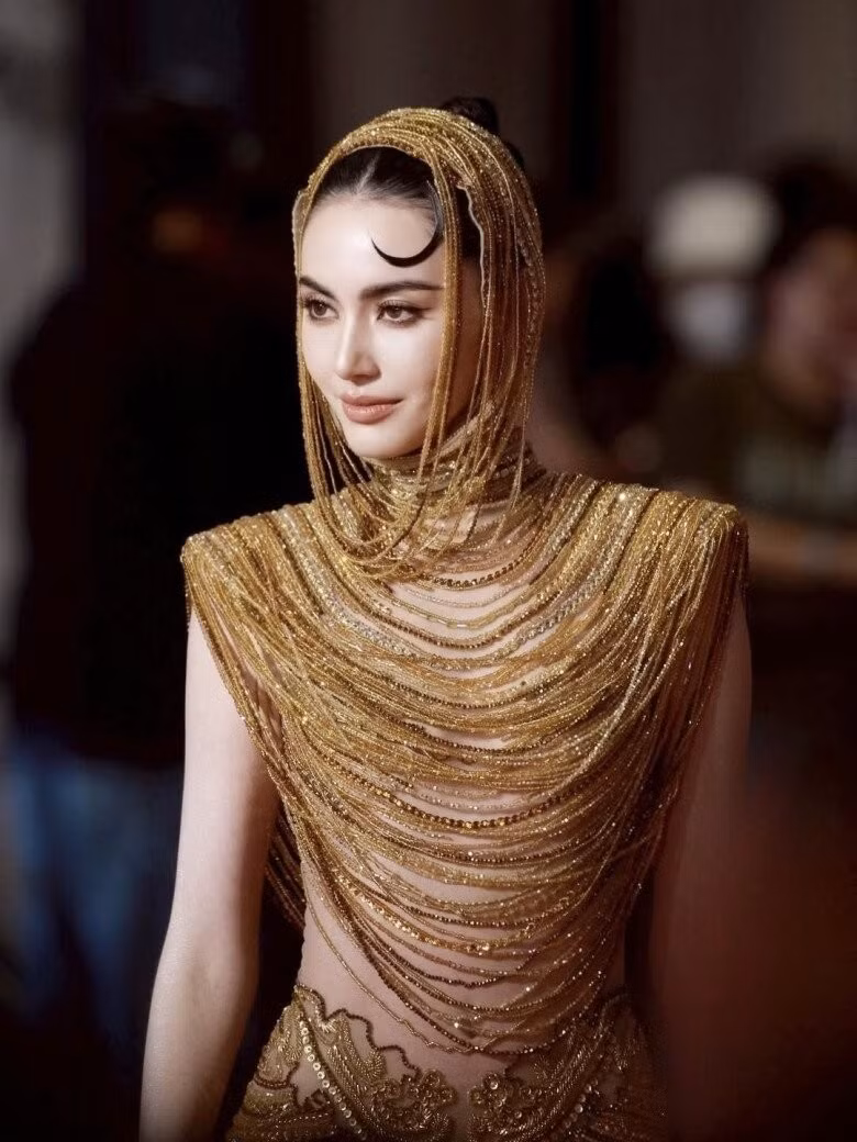 Điểm nhấn không thể bỏ qua chính là bộ váy ánh vàng ôm sát cơ thể, khoe trọn đường cong gợi cảm. Thiết kế với phần mũ trùm đầu liền mạch, những lớp xếp hạt cườm vàng óng ánh tựa những dải ánh sáng buông rủ xuống vai và thân váy.