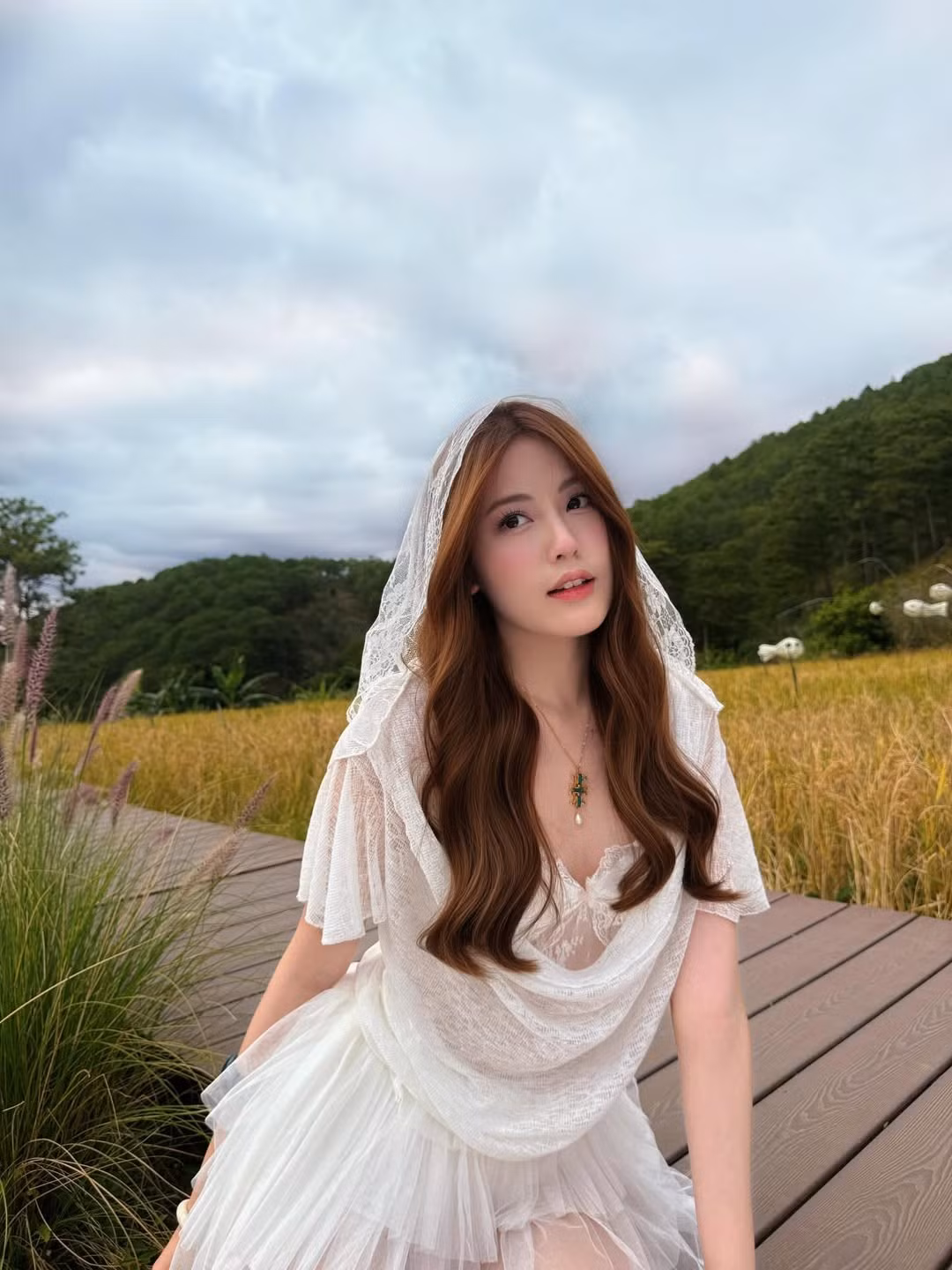 Trong loạt ảnh, Tăng Mỹ Hàn diện chiếc váy trắng mỏng nhẹ, được thiết kế nhiều lớp voan và ren tinh tế. Phom váy xòe nhẹ giúp tôn lên vóc dáng thanh mảnh, đồng thời mang lại cảm giác bay bổng, nữ tính. Điểm nhấn là phần cổ đổ mềm mại kết hợp tay áo sheer mỏng, vừa kín đáo vừa gợi nét mong manh. Chiếc khăn voan đội đầu càng làm tăng vẻ dịu dàng, hòa quyện hoàn hảo với không gian thiên nhiên xanh – vàng của cánh đồng lúa.