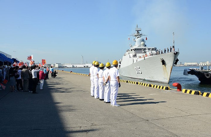 Các sĩ quan Lực lượng Phòng vệ trên biển Nhật Bản (JMSDF) xếp hàng chờ bắt dây neo để cố định tàu Trần Hưng Đạo vào cầu tàu. Phía sau là người dân đang vẫy quốc kỳ Việt Nam và Nhật Bản.