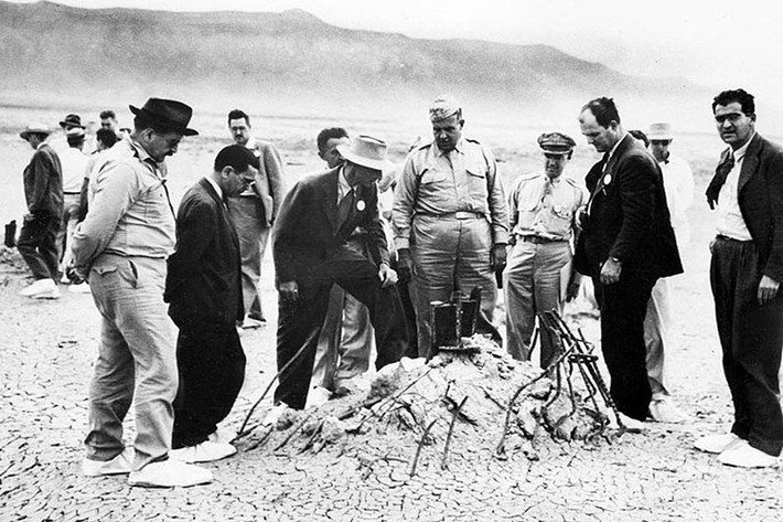 Hình ảnh Thiếu tướng Leslie Groves và Robert Oppenheimer tại tháp thử nghiệm Trinity sau khi vụ thử diễn ra vài tuần. Giày của họ được lót miếng bọc trắng đặc biệt để ngăn bụi phóng xạ dính vào giày. Nguồn ảnh: Wikimedia.