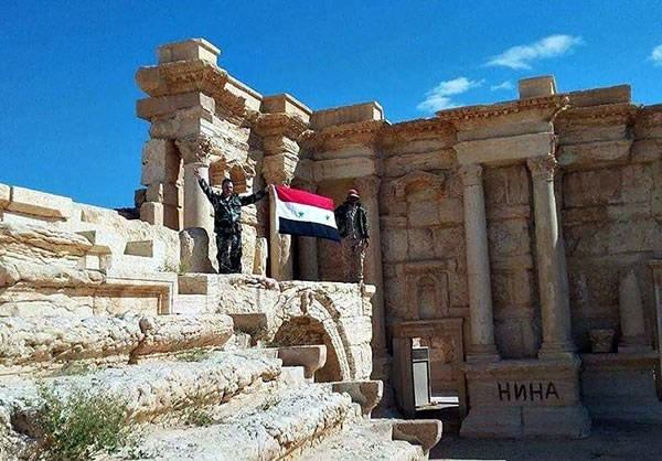 Hai binh sỹ Syria giơ cao cờ Syria tại một địa điểm lịch sử.