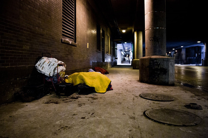 Một khu người vô gia cư thường tới lui ở đường Lower Wacker Drive, Chicago, nơi một người đang ngủ dưới nhiều lớp chăn ở cửa vào một tiệm thuốc. Trong khi người dân thành phố đang gấp rút mua hàng để chuẩn bị cho tình huống xấu nhất, người vô gia cư 60 tuổi này chỉ có thể đeo trên cổ tấm biển xin được giúp đỡ. Ông đã ngủ ngoài đường đêm trước đó, và cơ thể vẫn chưa thể ấm lên. Ông nói chưa biết sẽ ngủ ở đâu đêm nay. Ảnh: New York Times.