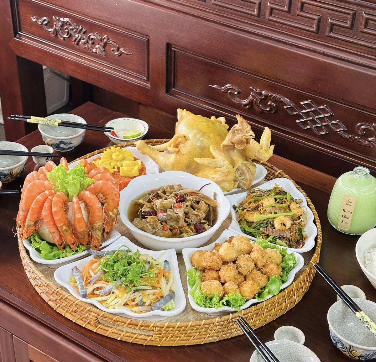 Mâm cỗ được bày gọn gàng trên mâm tre tròn, nổi bật với gà luộc nguyên con da vàng óng, đặt ở vị trí trung tâm. Xung quanh là tôm hấp xếp vòng, canh hầm thảo dược với thịt mềm và nước dùng trong, món xào thập cẩm, nem chiên giòn, chả cuốn và nộm rau củ thanh mát. Cách bày mâm mộc mạc nhưng đầy đủ món mặn món rau món nước, phù hợp với không khí cúng lễ ngày cuối năm. Ảnh Internet