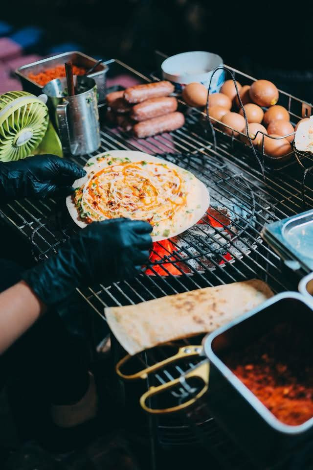 Nhắc đến chợ đêm Đà Lạt, bánh tráng nướng là món ăn đầu tiên được nhiều du khách lựa chọn. Những quầy bánh san sát nhau, luôn đỏ lửa suốt buổi tối. Từ chiếc bánh tráng mỏng, người bán khéo léo kết hợp trứng, mỡ hành, tép khô, xúc xích, phô mai… tạo nên món ăn giòn rụm, béo bùi, đậm đà. Mỗi quầy có cách nêm nếm riêng, mang đến hương vị khác biệt, đủ để thực khách ăn một lần là nhớ mãi. Ảnh Nguyên Phú