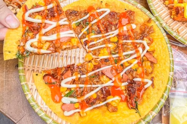 Chỉ với chiếc bếp than nhỏ, vài lát bánh tráng và topping như trứng gà, xúc xích, hành phi, phô mai..., các cô bán hàng vỉa hè đã tạo nên những “chiếc pizza Việt” thơm lừng, giòn rụm. Đây là món ăn vặt cực kỳ hút giới trẻ, giá dao động từ 10.000 – 20.000 đồng/chiếc, đặc biệt phổ biến ở khu vực gần Quảng trường 2/4. Ảnh Cookpad