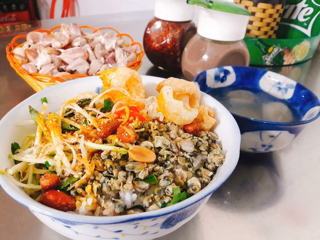 Nếu du khách đang muốn trải nghiệm vị dân dã đặc trưng của Huế, hãy ghé quán cơm hến Hoa Đông ở đường Ưng Bình. Chỉ với 10.000 – 20.000 đồng, bạn sẽ có một đĩa cơm hến đầy đủ hến xào, tóp mỡ giòn tan, rau sống thái nhỏ, đậu phộng và mắm ruốc cay – tất cả trộn đều với nước hến nóng, tạo nên hương vị cay nồng khó quên. Ảnh MIA